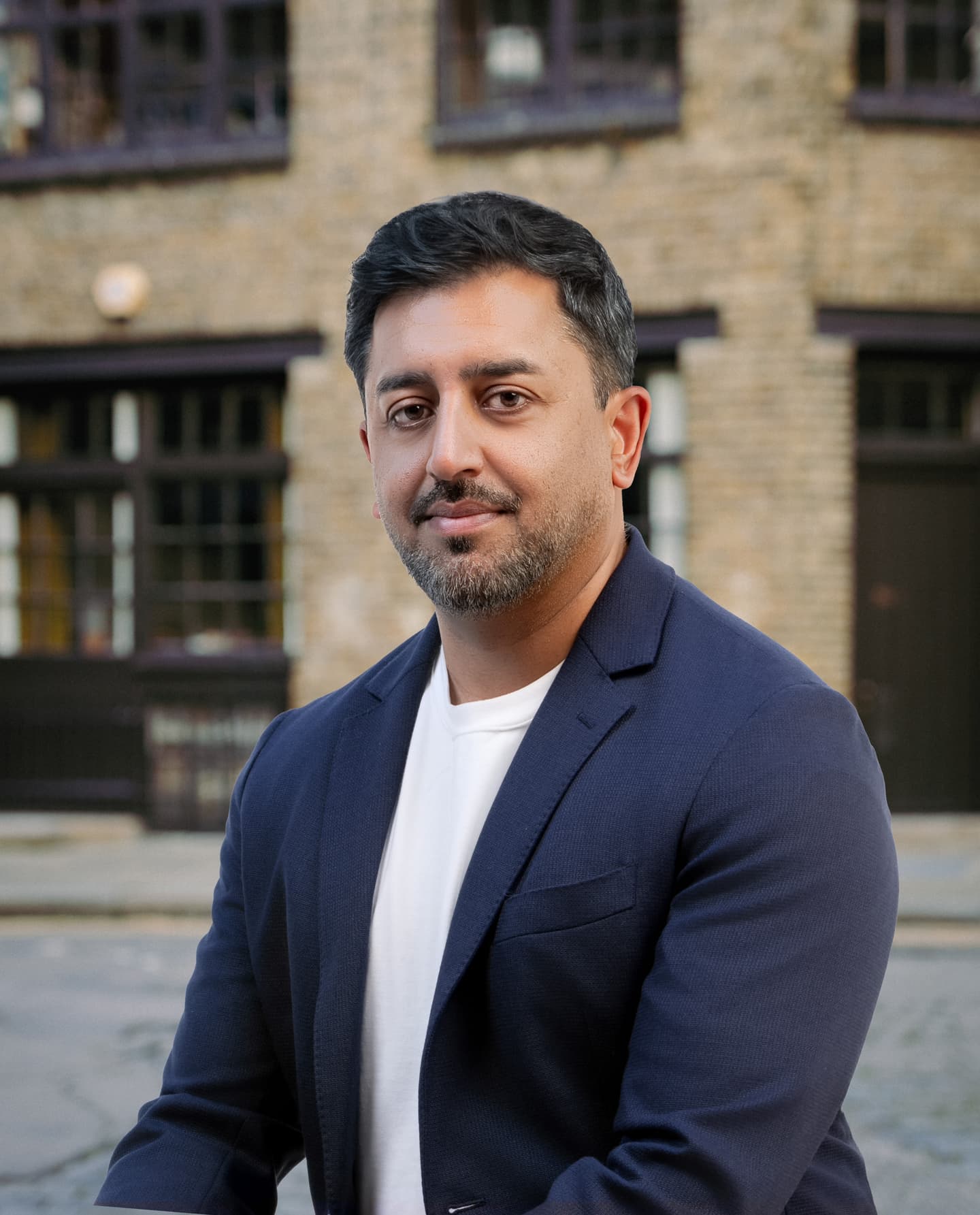 A man with short dark hair and a trimmed beard wears a navy blazer over a white shirt, sitting outdoors in front of a brick building.