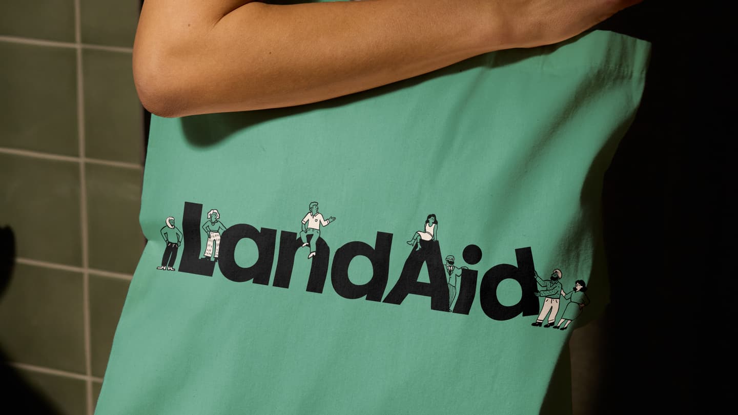A person holds a green tote bag with the word LandAid printed in large black letters, featuring small illustrated people around the text.