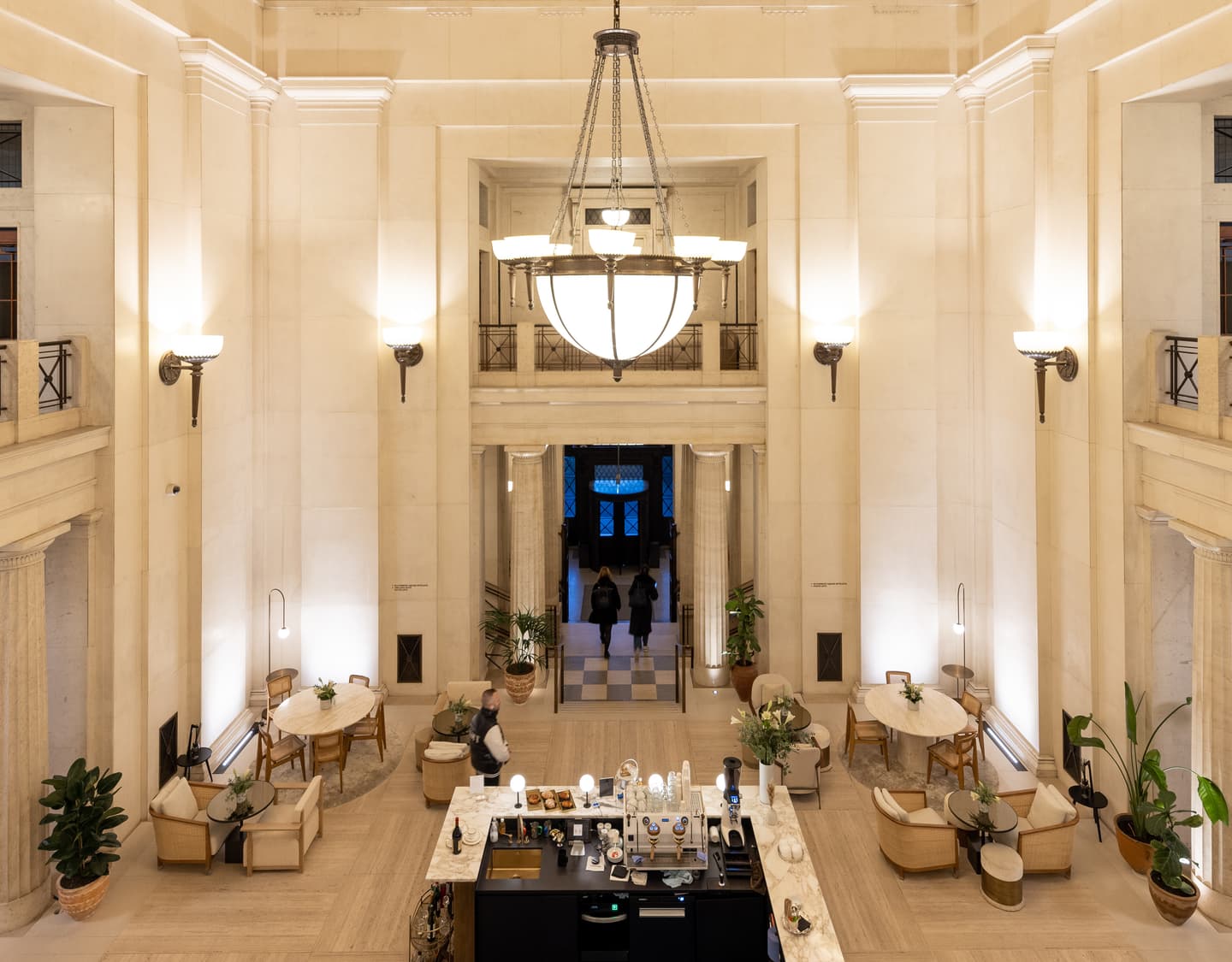 A spacious, elegant lobby with high ceilings, neutral walls, a central coffee bar, seating areas, potted plants, and people entering through double doors in the background.