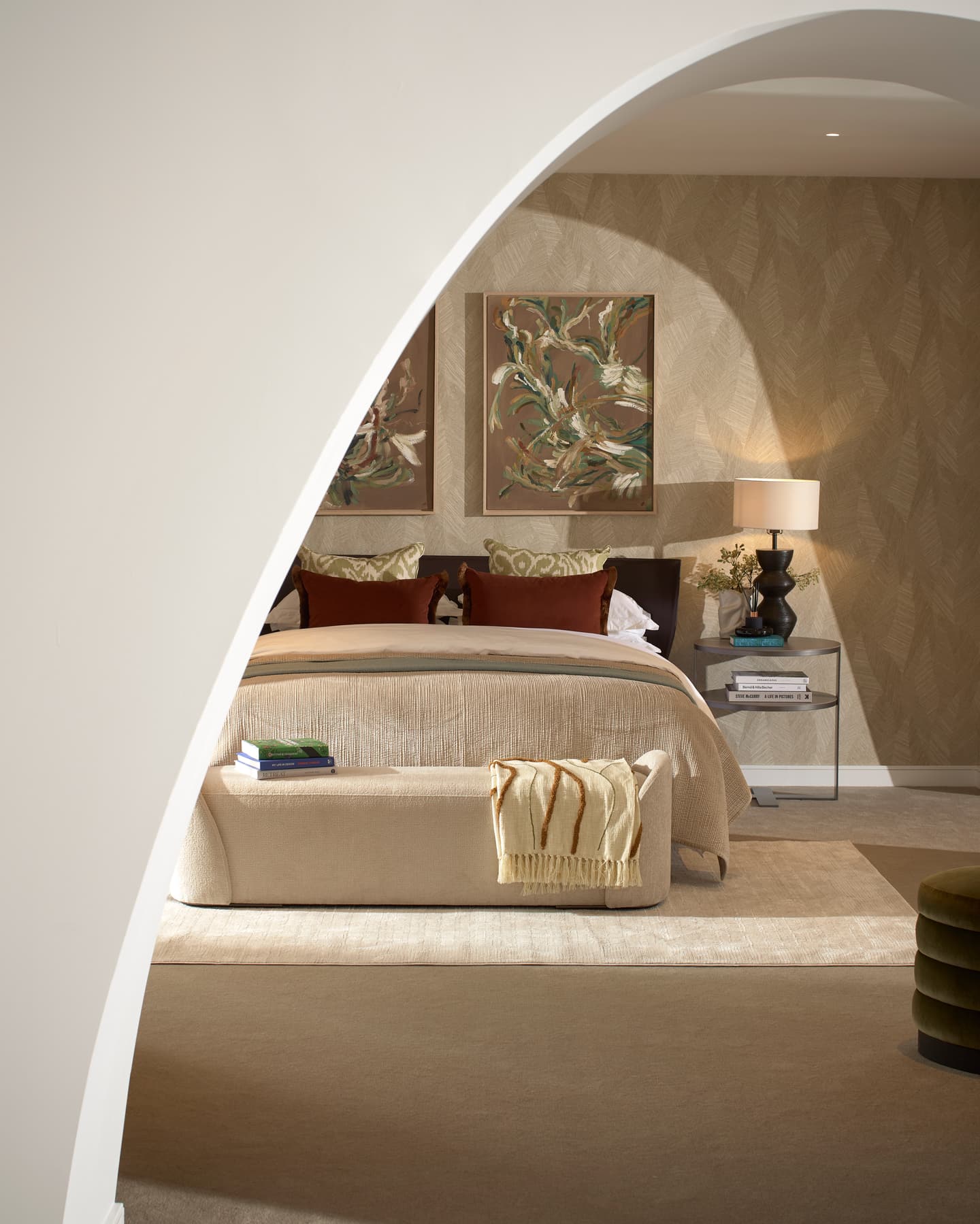 A modern bedroom with a beige colour scheme, seen through an arched doorway, features a bed, ottoman, two paintings, a side table with a lamp, and neatly arranged decor.