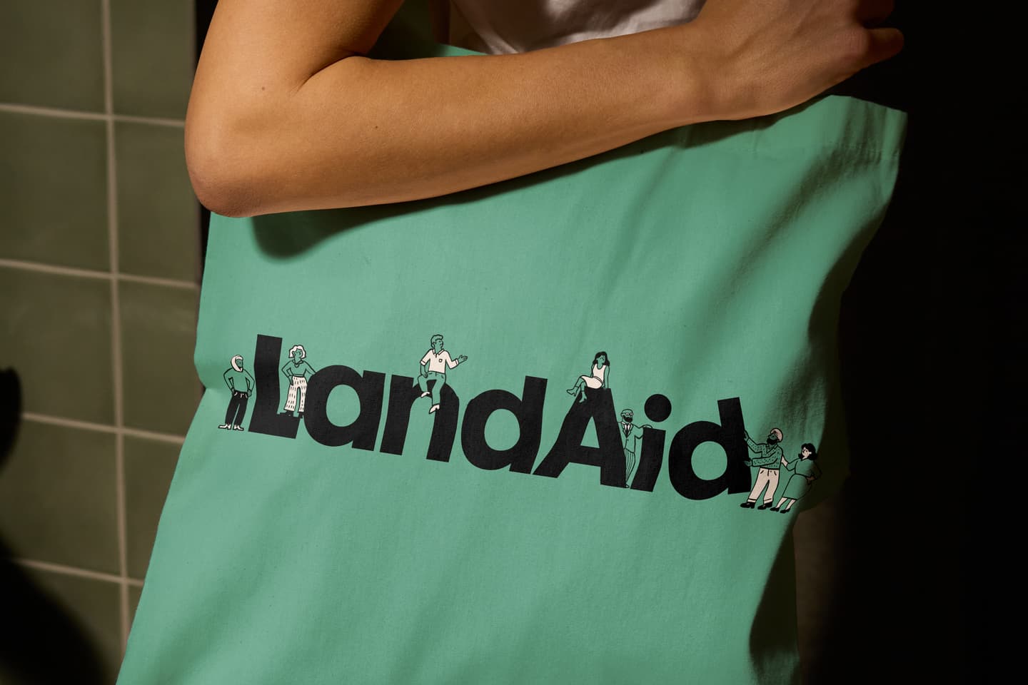 A person holds a green tote bag with the word LandAid printed in large letters, each letter illustrated with small cartoon figures.
