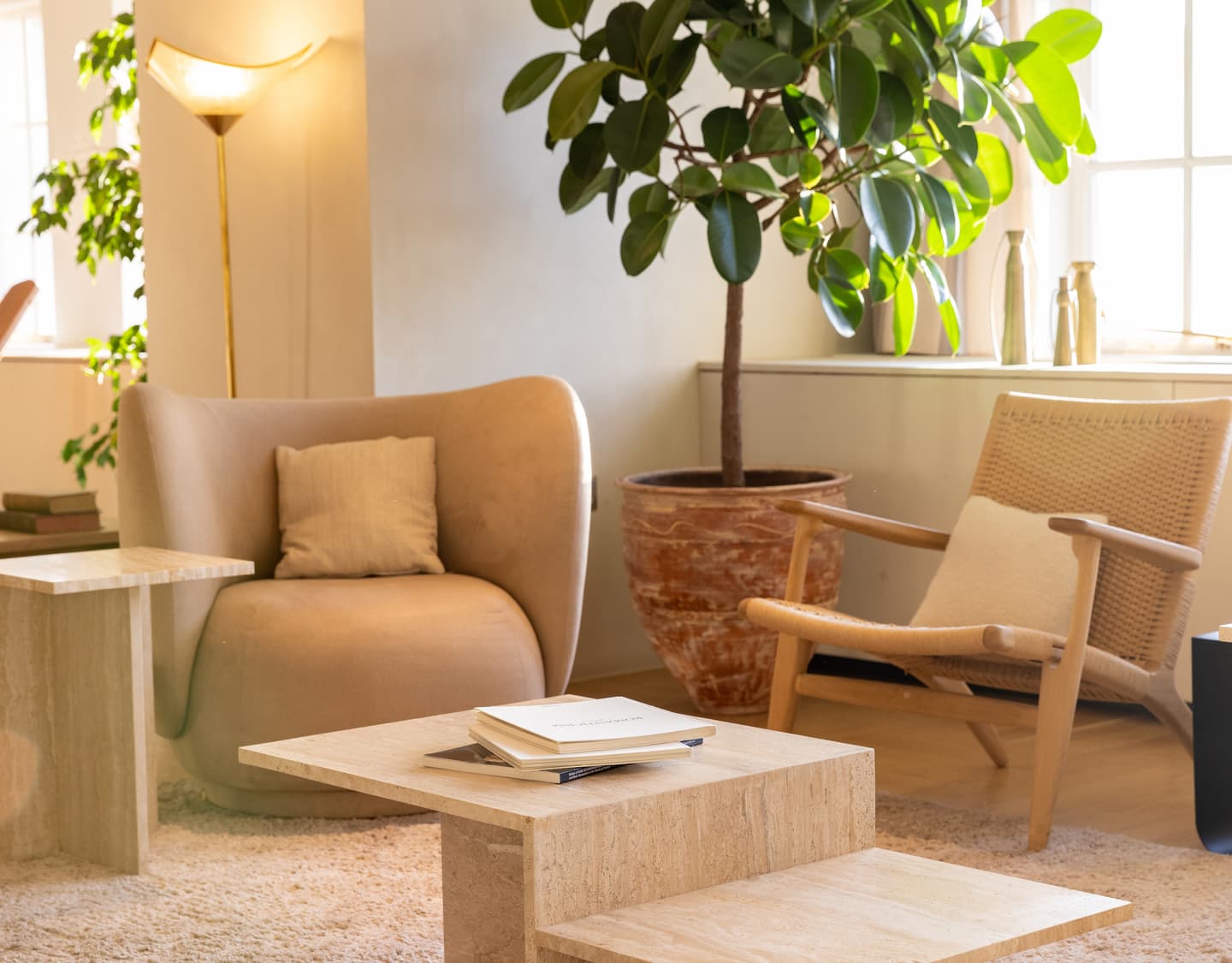 A cosy living room with a beige armchair, a woven chair, a potted plant, light wood tables, and a book, all illuminated by natural light from large windows.