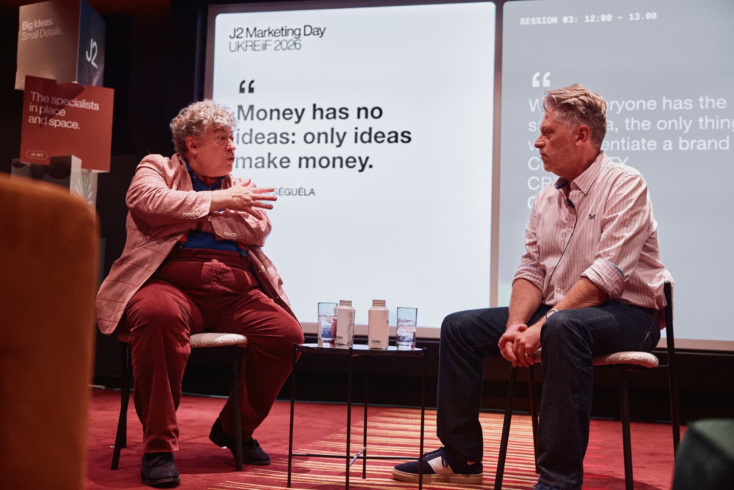 Two men sit and talk on stage at a marketing event, with a projection behind them showing the quote Money has no ideas: only ideas make money.