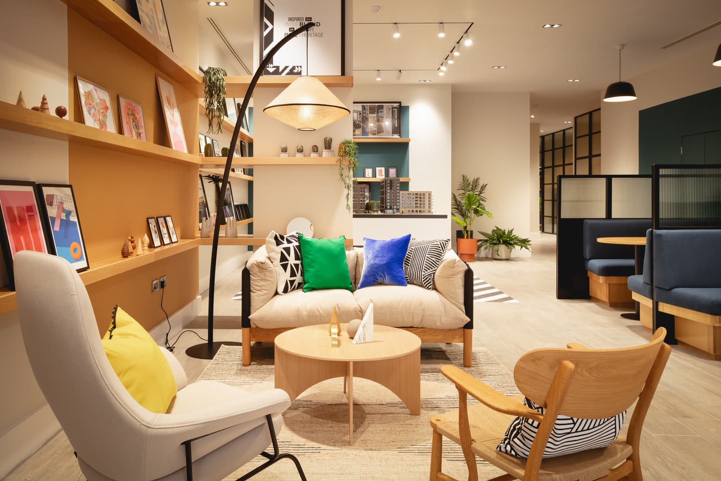 Modern living room with a beige sofa, colourful cushions, wooden chairs, a round coffee table, wall shelves with art and decor, and bright, warm lighting.