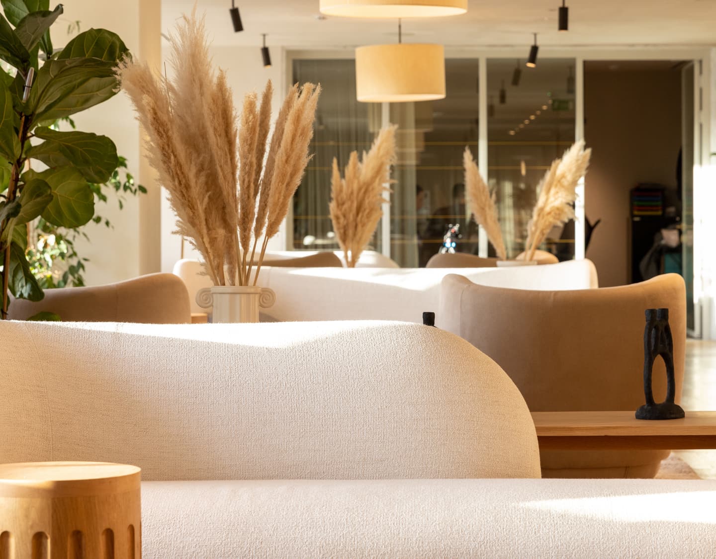 Modern lounge area with white curved sofas, pampas grass in vases, wooden tables, large plant, and neutral lighting in a bright, open space.
