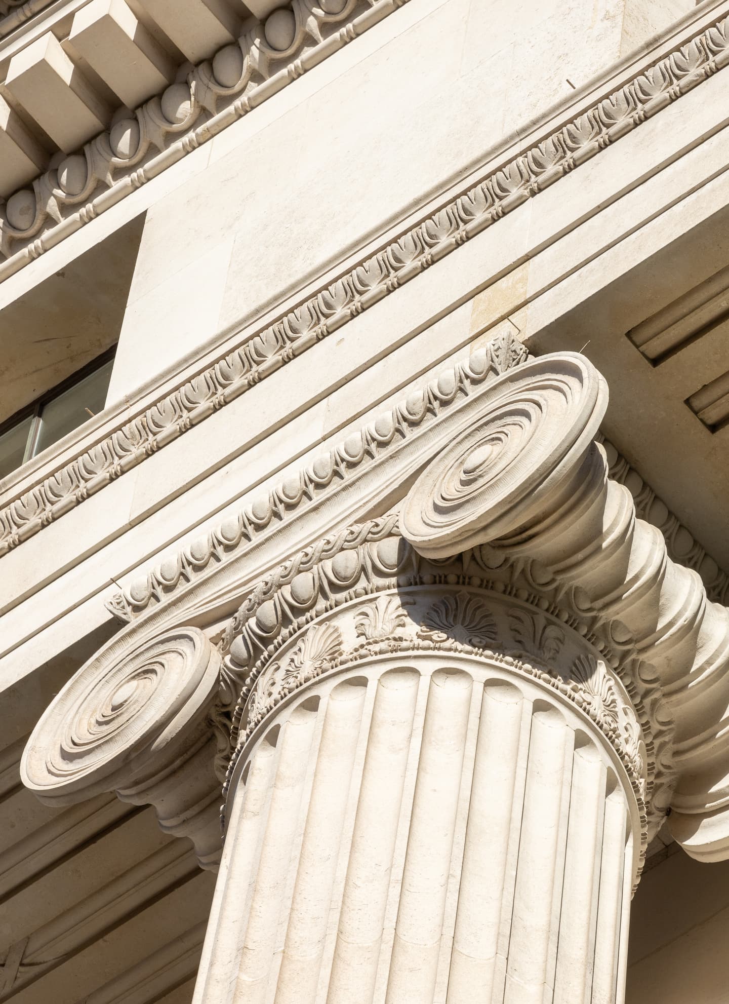 Close-up view of an ornate Ionic column with fluted shaft and volute capitals, supporting a detailed entablature on a classical stone building.