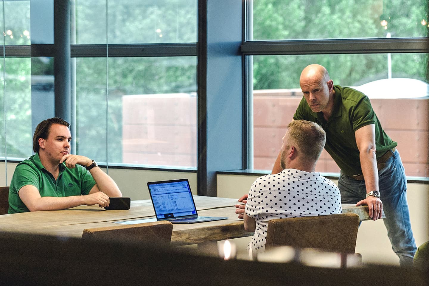 Three men have a discussion around a table in a modern office; one man is standing, two are seated, with a laptop open displaying a spreadsheet.