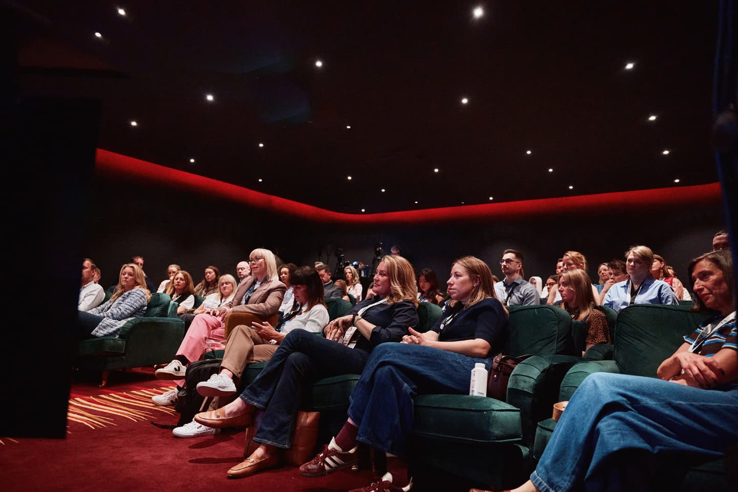 A group of people sit in rows of green chairs in a dimly lit room, attentively watching something off camera.