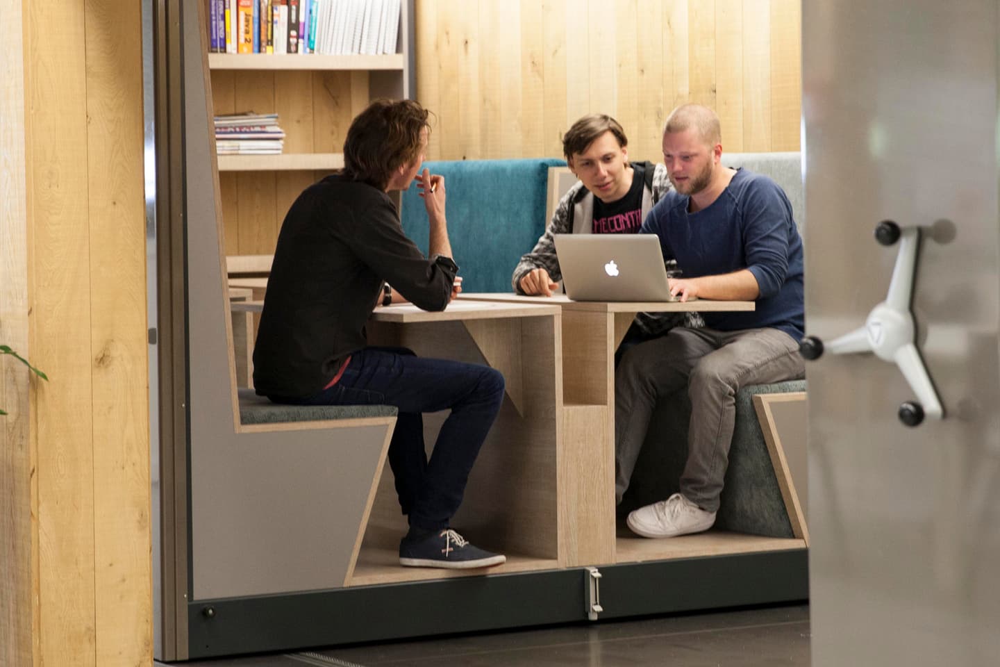 Three people sit in a wooden booth, collaborating and looking at a laptop. Books are visible on shelves above them.