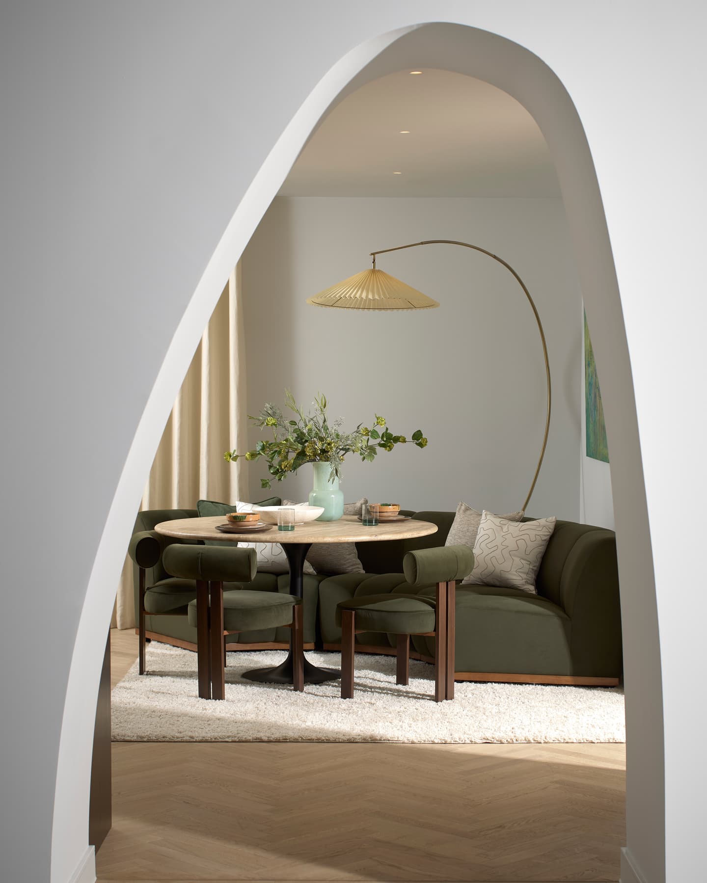 A modern dining and living area viewed through an arched doorway, featuring a round table, green chairs, a curved sofa, a pendant lamp, and a vase with flowers.