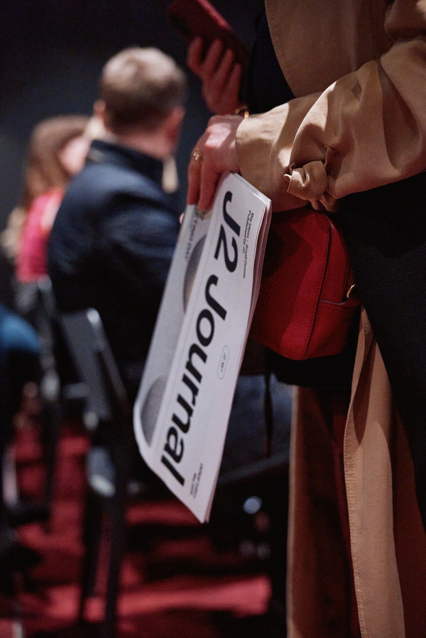 Person holding a folded J2 Journal whilst standing among seated people at an indoor event.