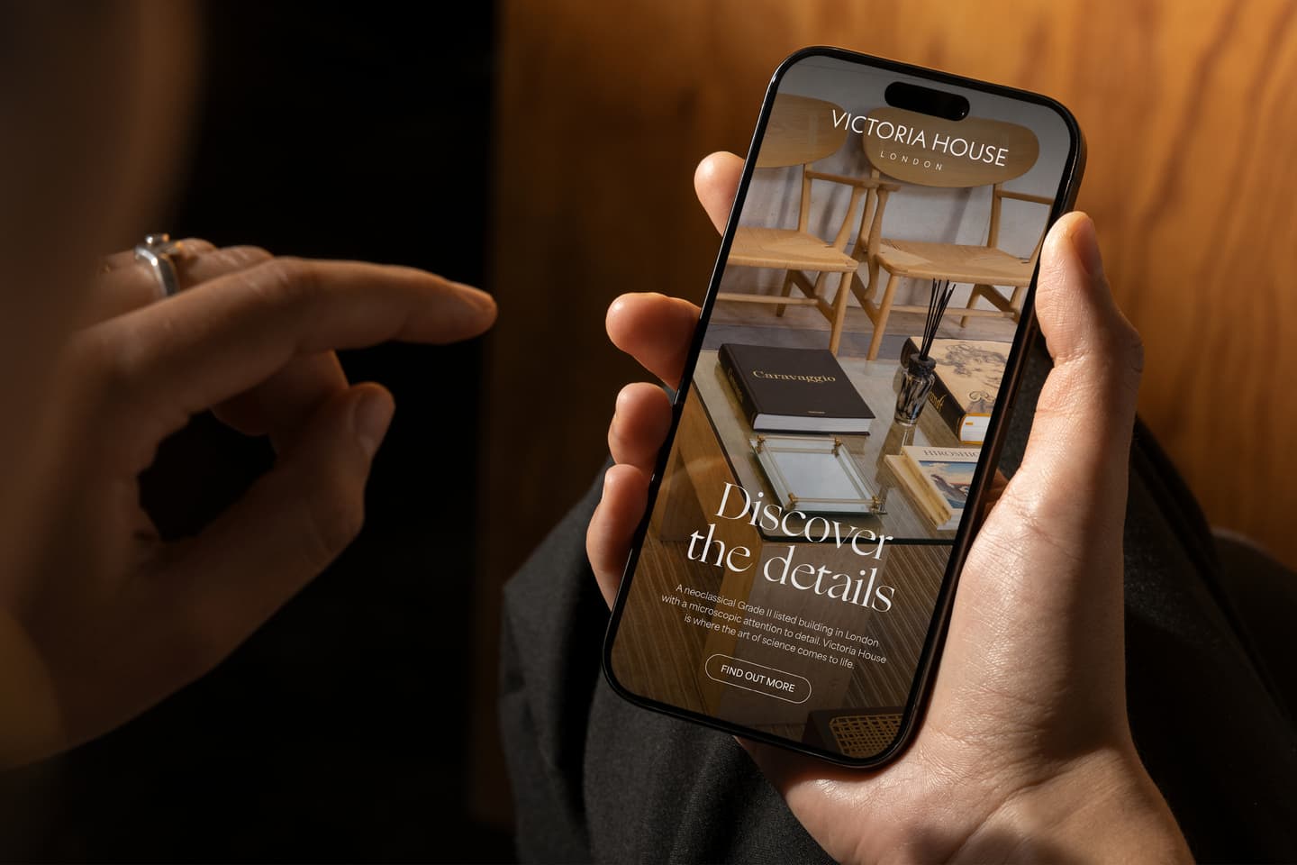 A person holds a mobile phone displaying a website for Victoria House London with the text Discover the details and a button labelled Find Out More.