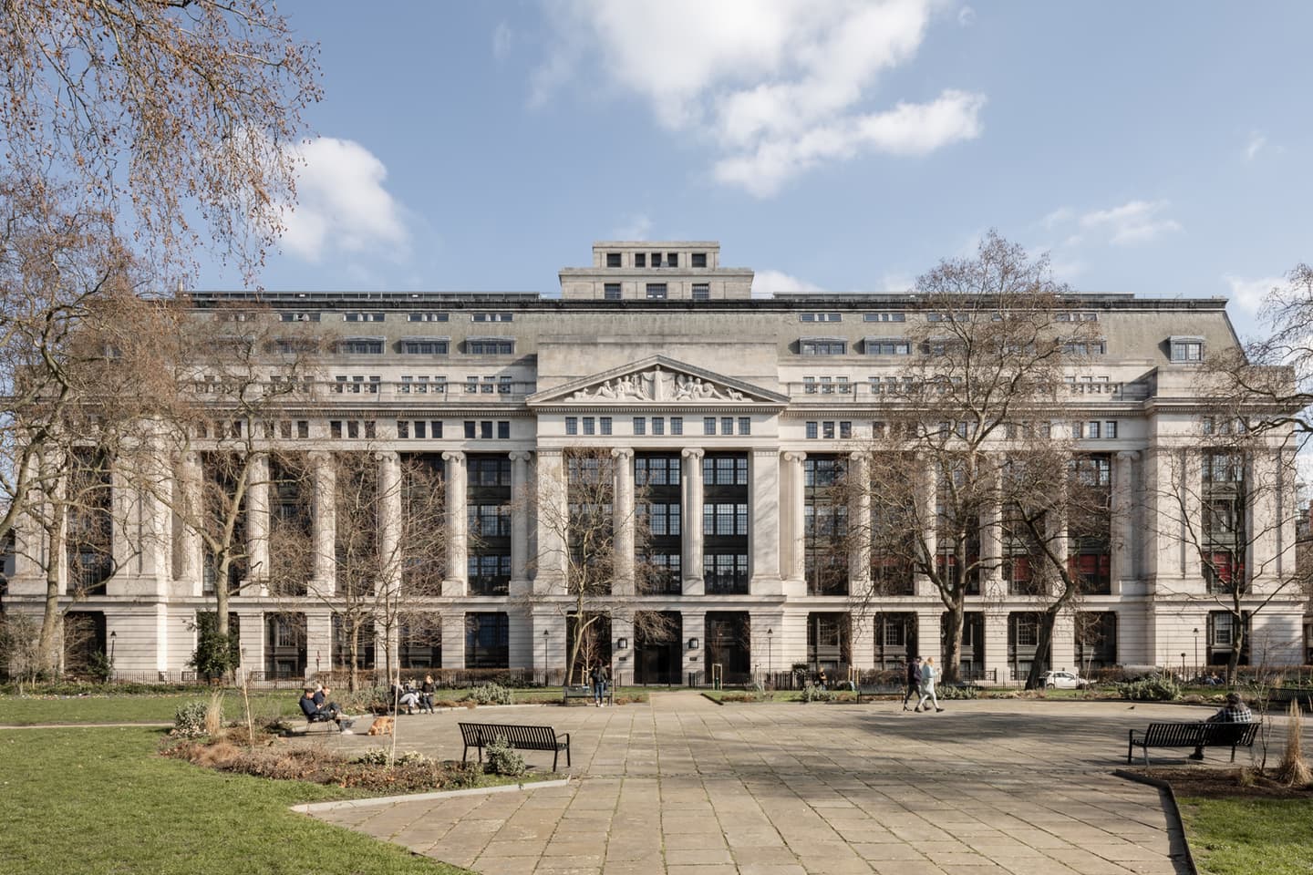 A large neoclassical building with columns and multiple windows stands behind a park with trees, benches, and people sitting or walking.