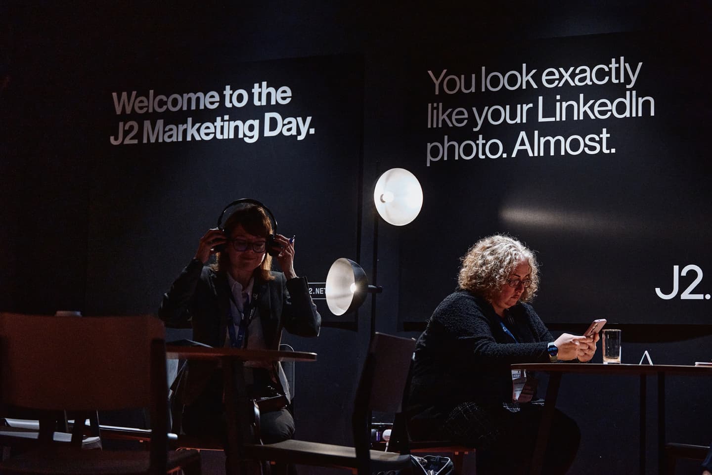 Two people sit at separate tables in a dimly lit room with signs on the wall that read Welcome to the J2 Marketing Day and You look exactly like your LinkedIn photo. Almost.