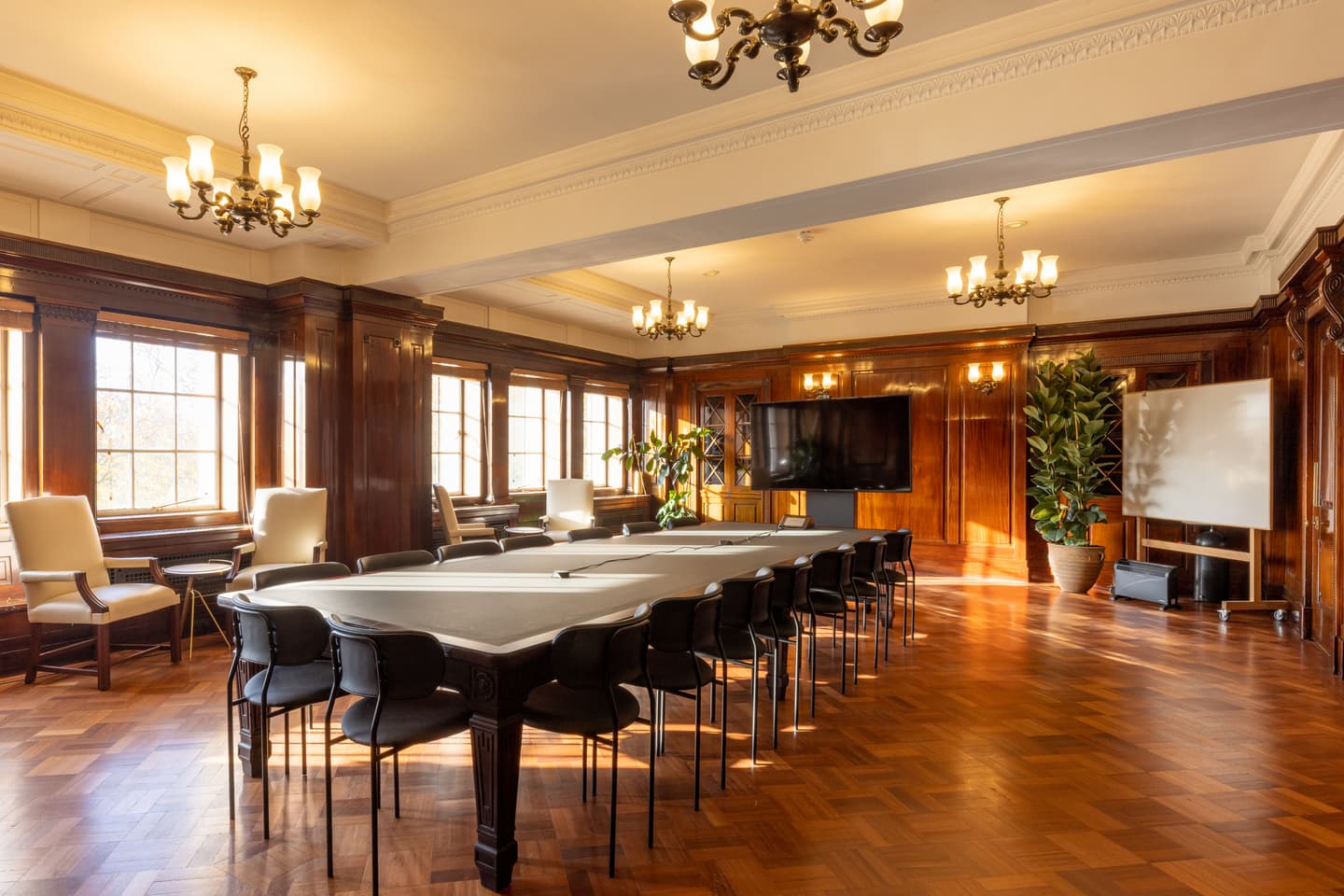 Spacious conference room with a long table, black chairs, large windows, wood panelling, chandeliers, a television, whiteboard, armchairs, and indoor plants.