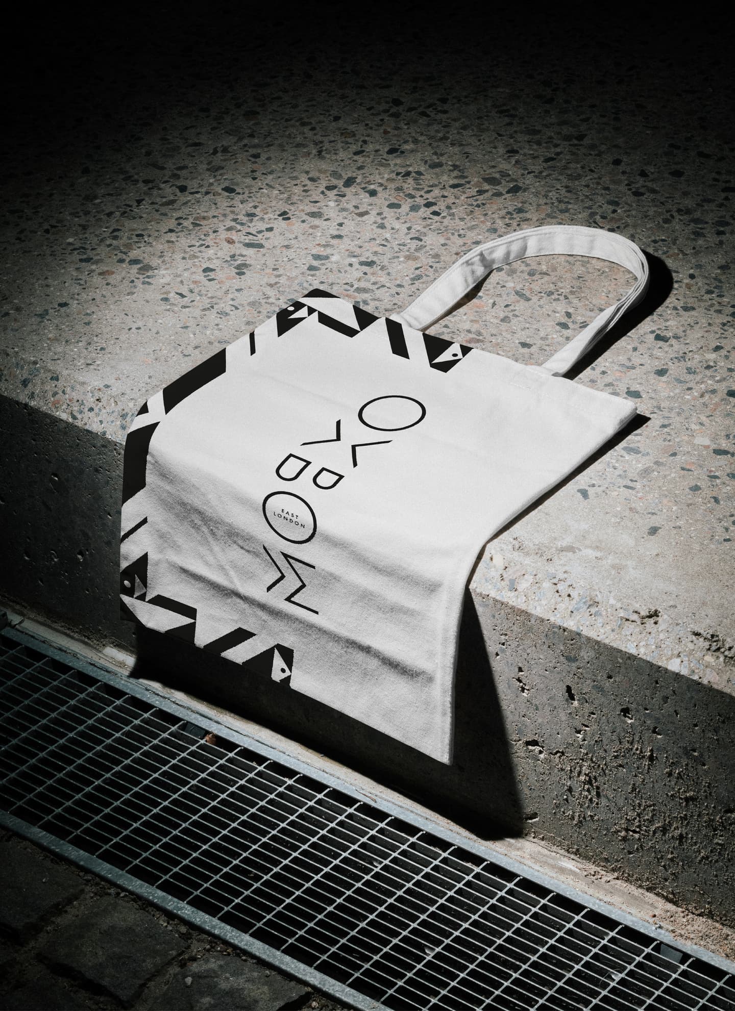 A white tote bag with black geometric designs and illegible text lies on a concrete ledge next to a metal grating.