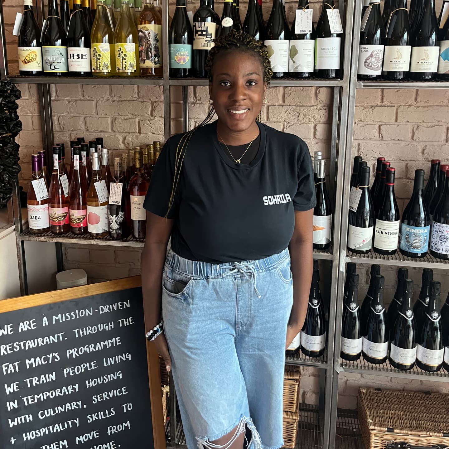 A woman stands smiling in front of shelves filled with wine bottles, next to a blackboard describing the mission of a restaurant programme.