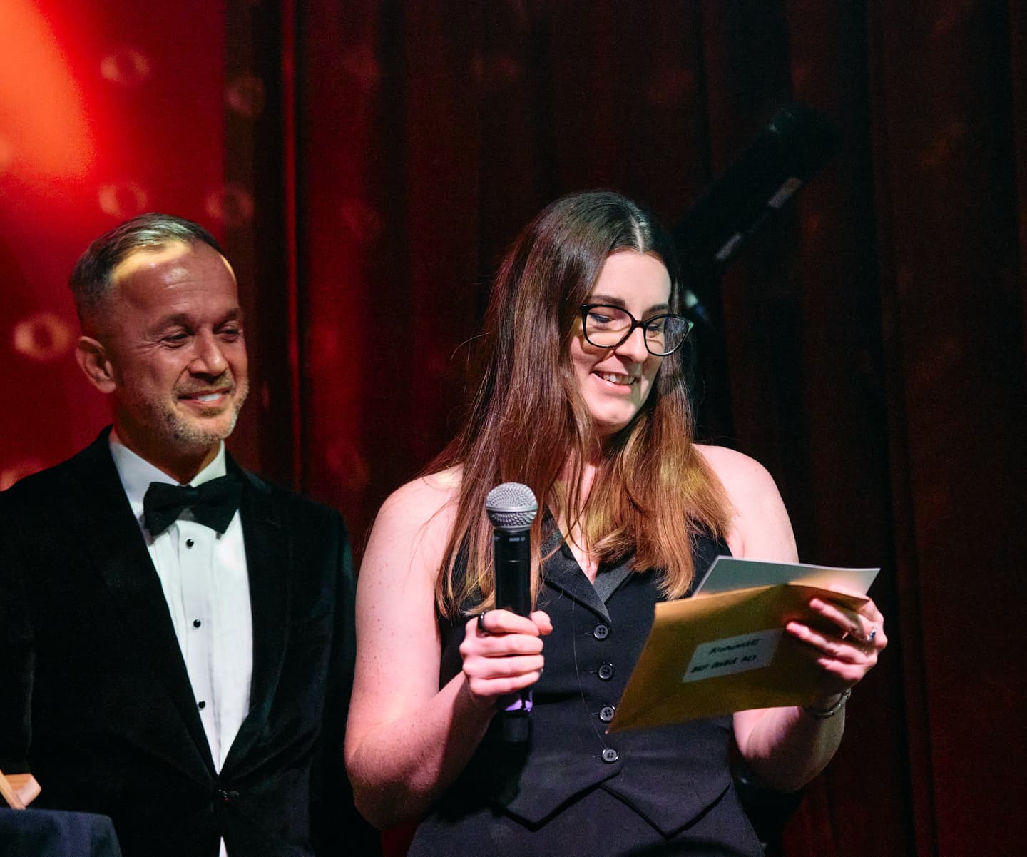 A woman holding a microphone and an envelope reads from a piece of paper on stage, while a man in a dinner jacket stands beside her, both appearing at an event with a red curtain backdrop.