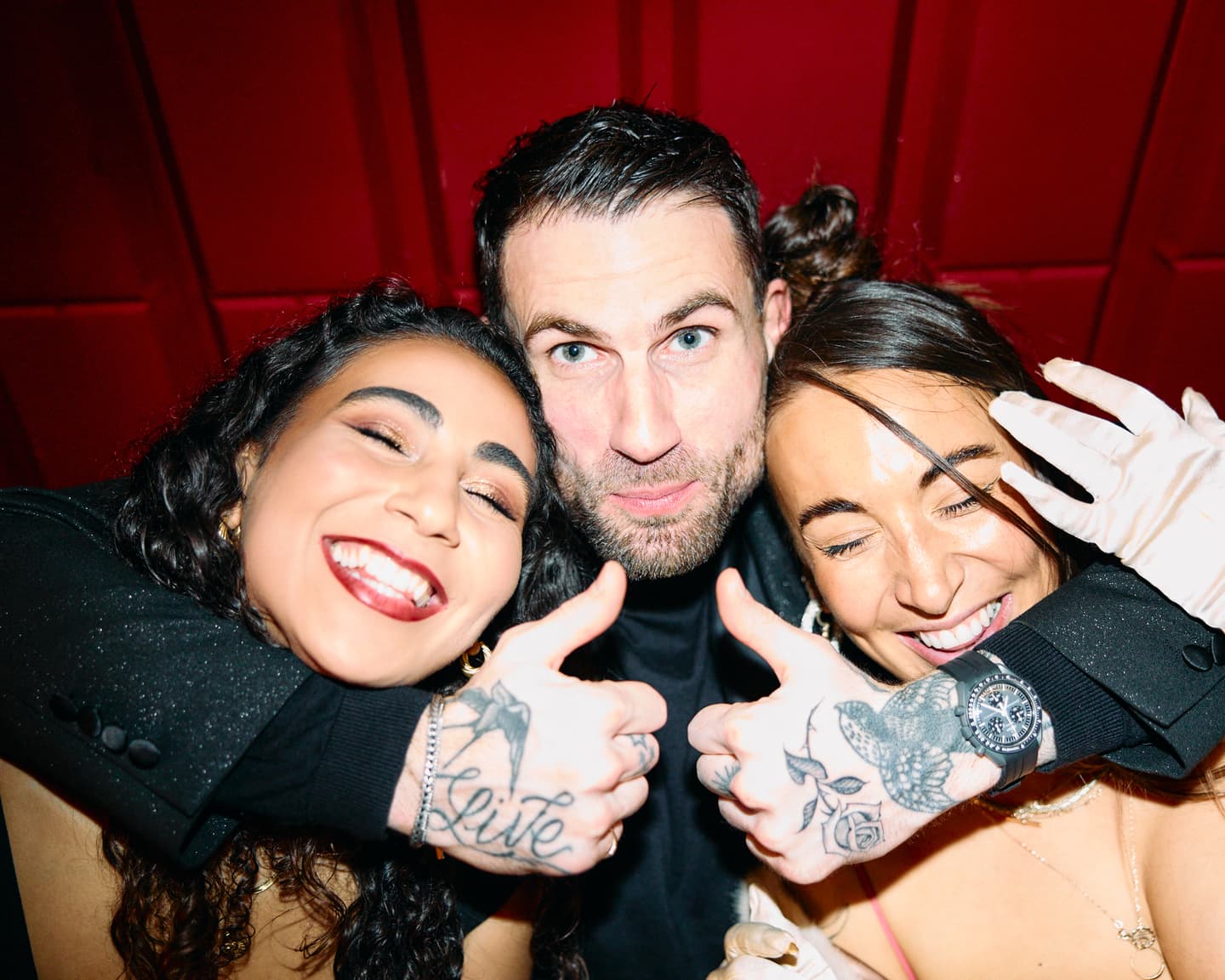 Three people pose closely together in front of a red background, smiling and giving thumbs up. Visible tattoos are on the hands of the person in the middle.