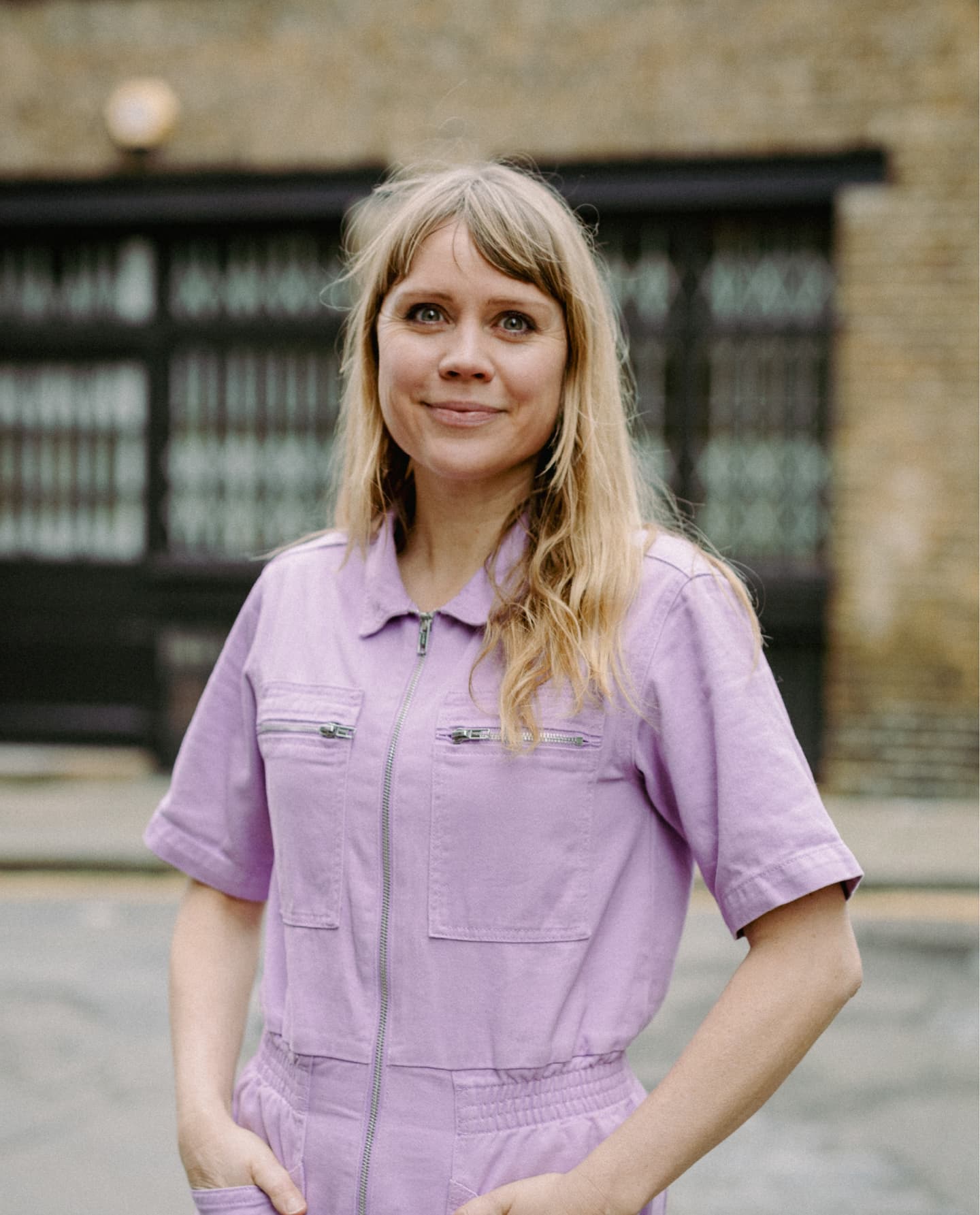 A person with long blonde hair stands outdoors wearing a light purple zip-up playsuit, with a brick building and metal gates in the background.