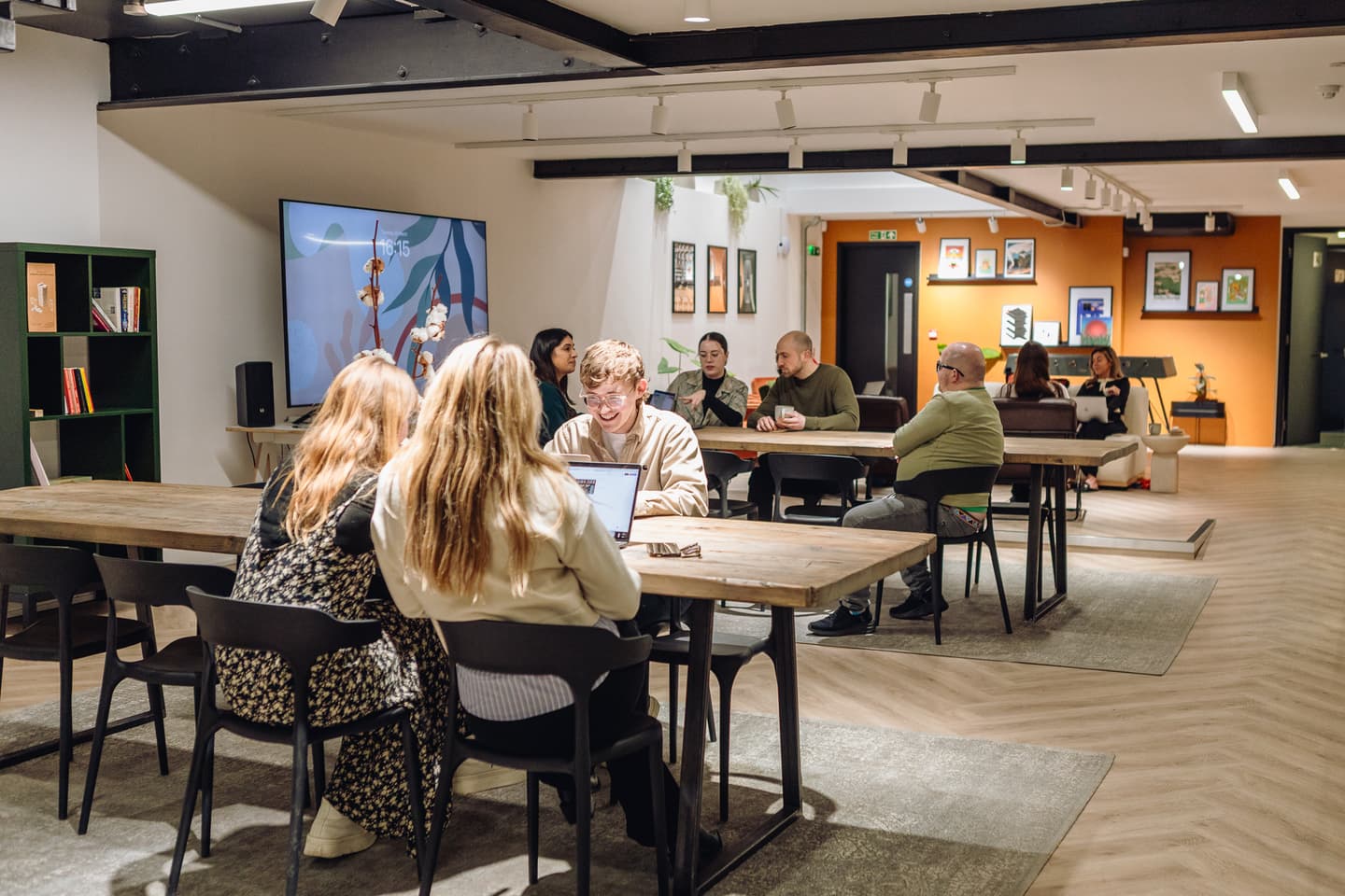 People sit at wooden tables in a modern, open workspace, talking, using laptops, and watching a large wall-mounted screen. Framed art decorates the orange and white walls.