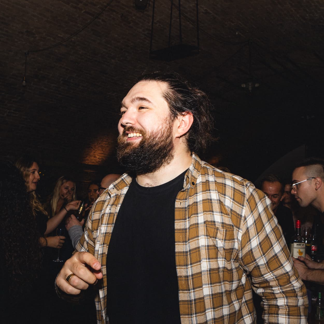 A bearded man in a checked shirt smiles and gestures while standing in a dimly lit, crowded indoor party setting.