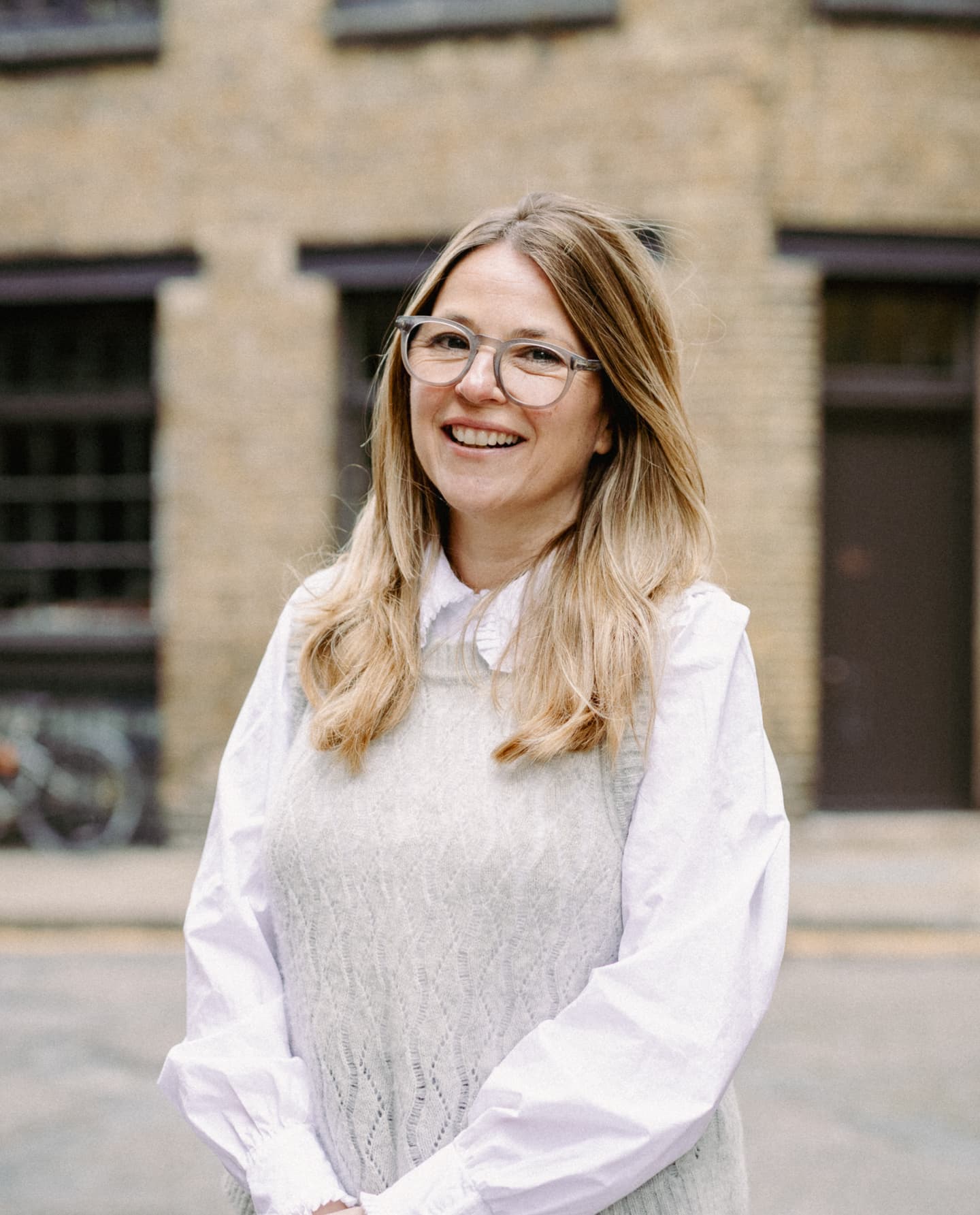 A woman with long blonde hair and glasses stands outdoors in front of a brick building, wearing a white blouse and light grey sleeveless jumper, smiling at the camera.