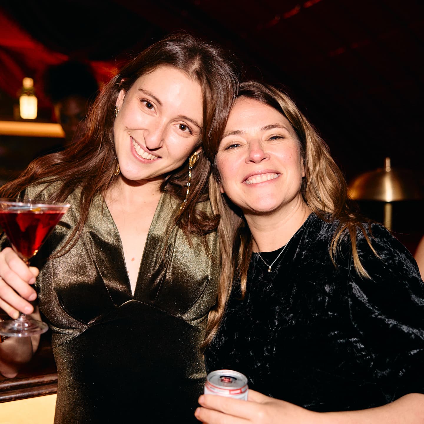 Two women smiling at the camera in a dimly lit indoor setting; one holds a cocktail and the other holds a tin.