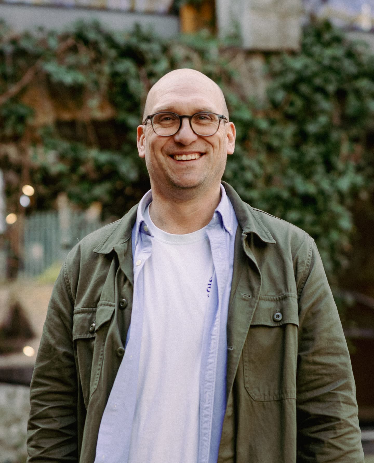 A man with glasses, wearing a white T-shirt, light blue shirt, and green jacket, stands outdoors in front of greenery, smiling at the camera.
