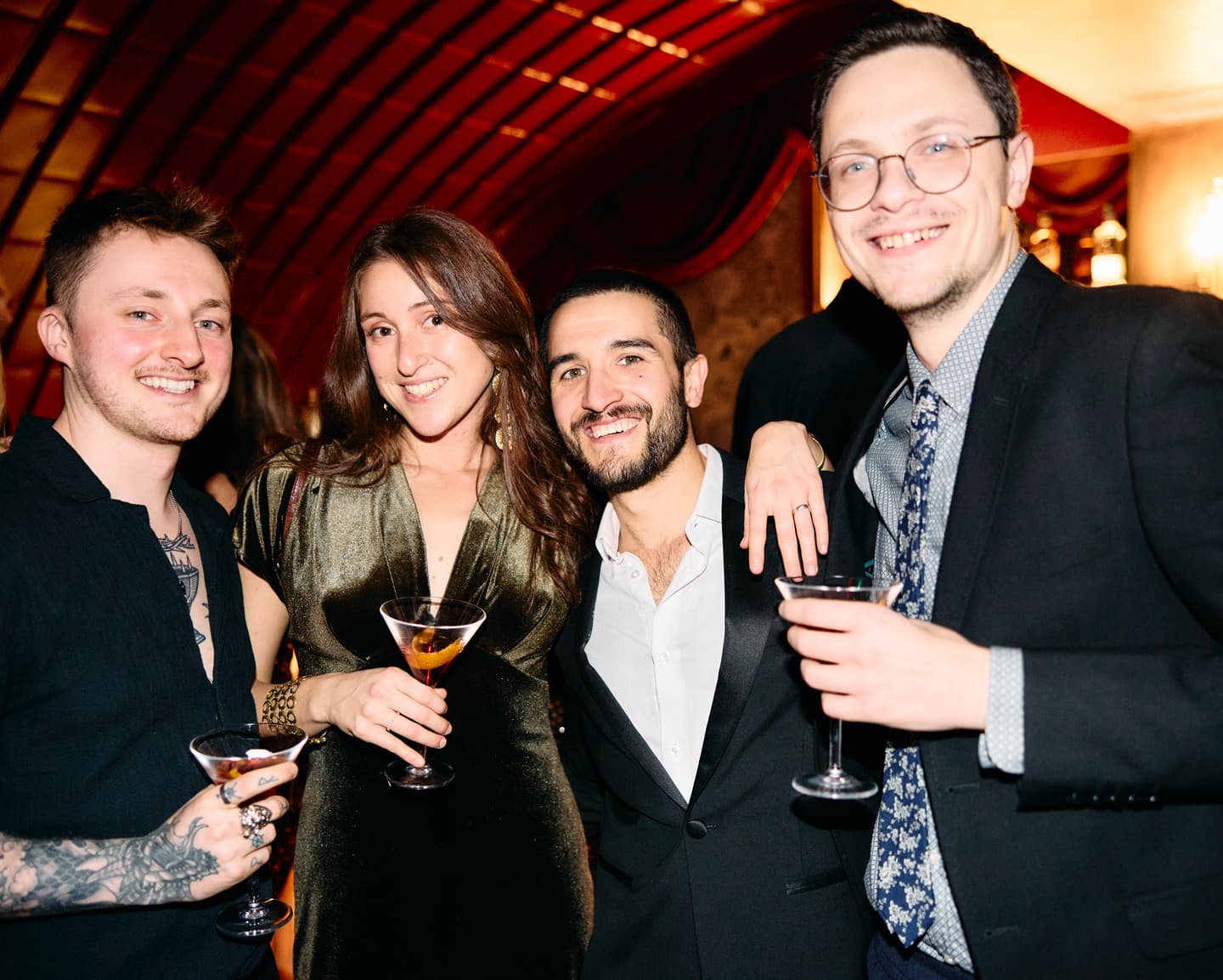 Four people dressed smartly pose together at an indoor event, each holding a cocktail glass and smiling at the camera.