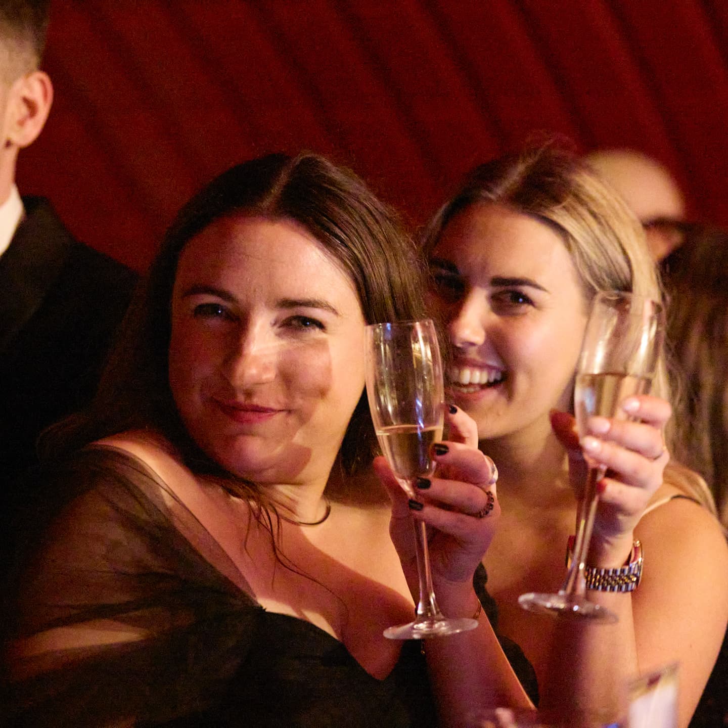 Two women dressed formally hold champagne glasses and smile at the camera at what appears to be a celebratory or social event.