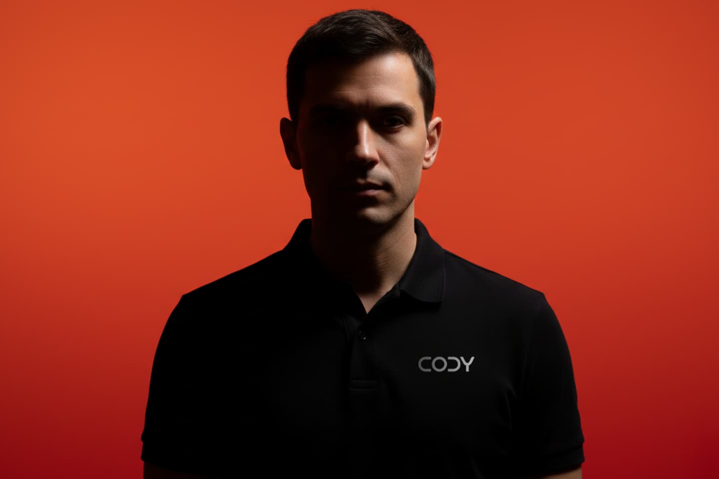 A man in a black polo shirt with CODY on it stands against a solid orange background, partially in shadow.