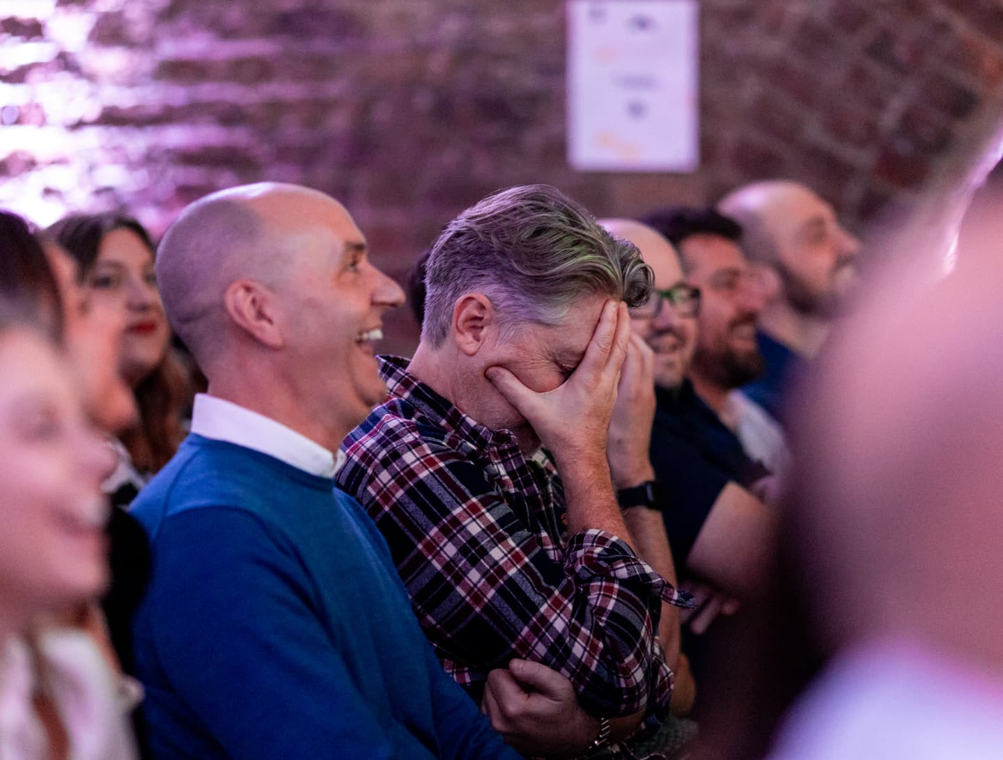 Several people are seated in an audience; one man in a checked shirt covers his face with his hand while others around him laugh and smile.