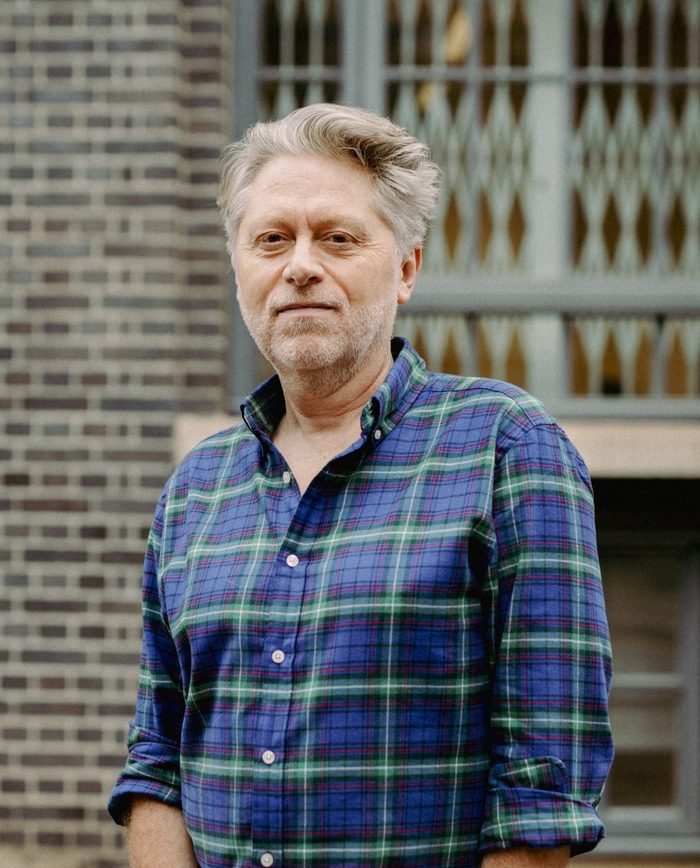 A man with grey hair and a beard wearing a blue and green checked shirt stands in front of a brick building.