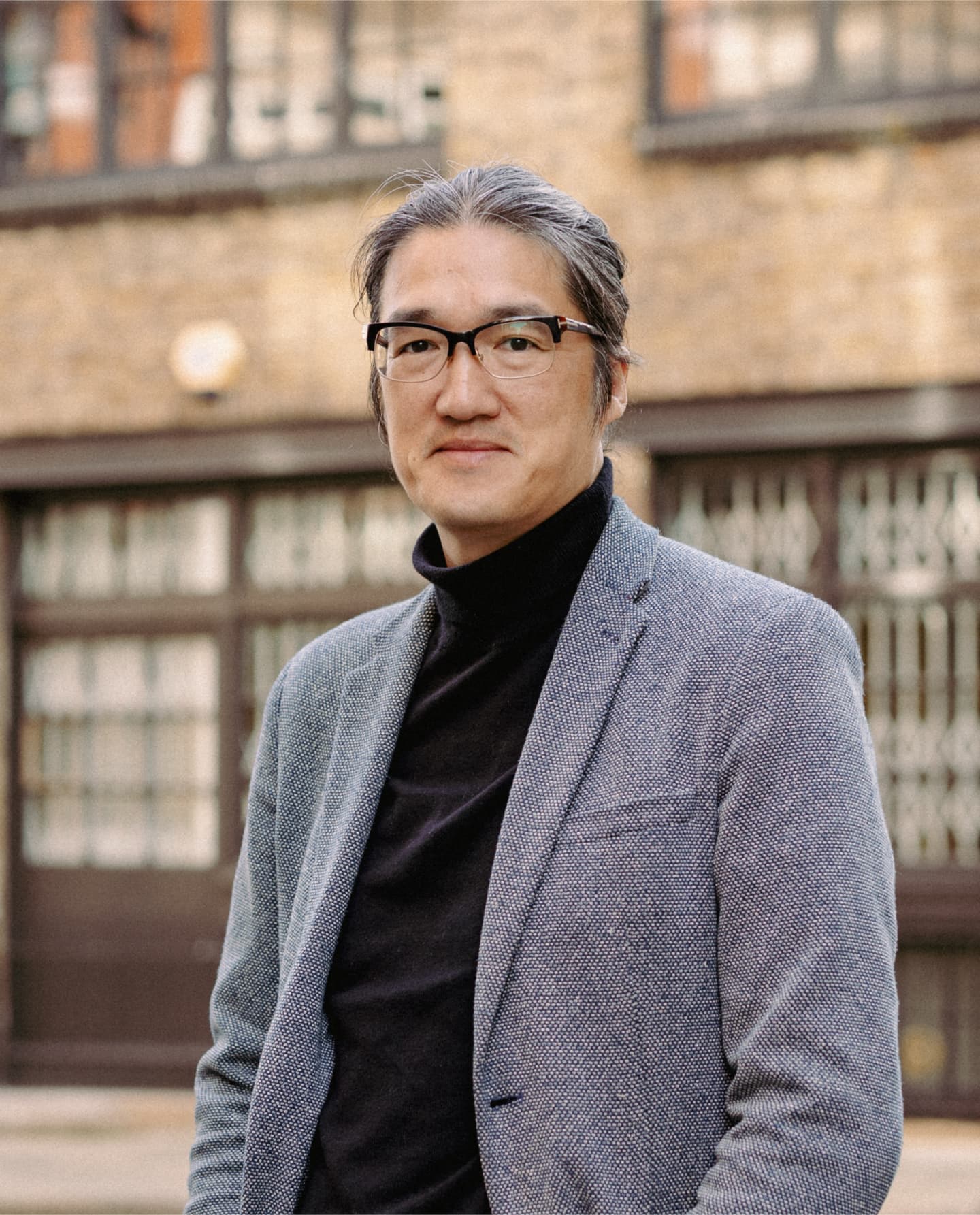 A person with glasses and grey hair wearing a grey blazer and black polo neck stands outdoors in front of a brick building with large windows.